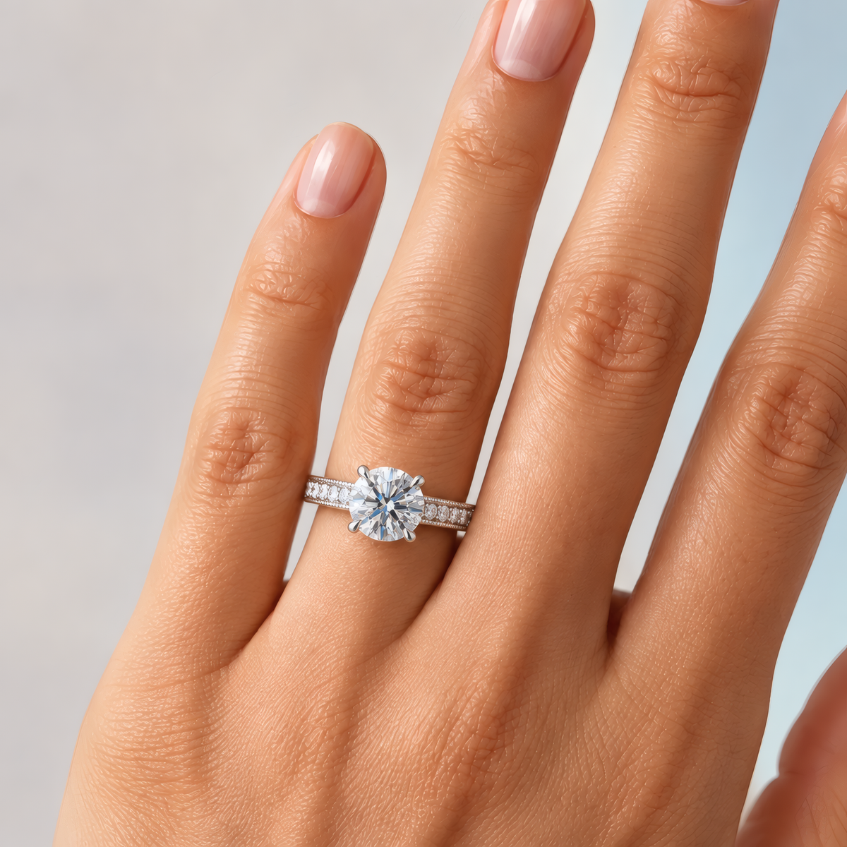 Delicate woman's left hand with a round solitaire engagement ring occupying the ring finger. Small round diamonds and milgrain cover the shank.