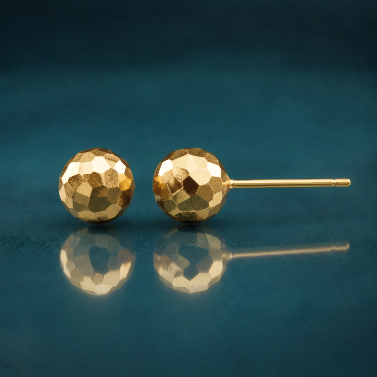 7mm faceted diamond-cut puffed ball stud earrings in 14k yellow gold, on a reflective teal surface.