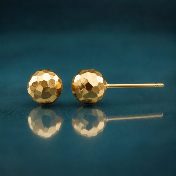 7mm faceted diamond-cut puffed ball stud earrings in 14k yellow gold, on a reflective teal surface.