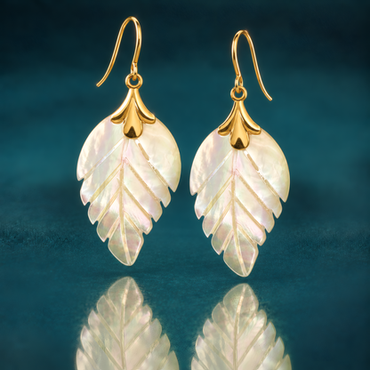 14k yellow gold dangle earrings with carved mother of pearl leaves on French hooks, on a reflective teal surface.