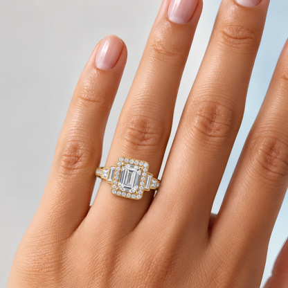Unique gold halo engagement ring with an emerald-shaped center and 2 baguette diamonds on each side of the halo, moving down the shank. The ring is worn on the ring finger of a delicate woman's left hand.