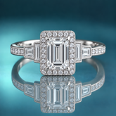 Unique white gold halo engagement ring with an emerald-shaped center and 2 baguette diamonds on each side of the halo, moving down the shank. The rest of the shank is set with round diamonds via prong in channel.
