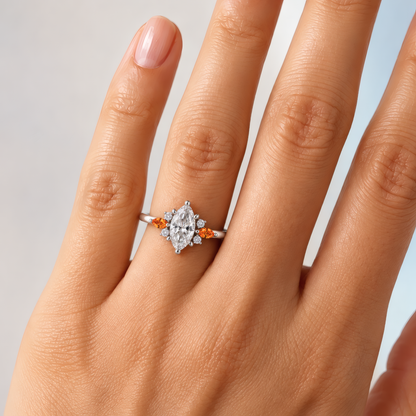 A delicate woman's left ring finger wearing a white gold engagement ring with a striking marquise center diamond, flanked on each side by a fiery amber marquise accent and a cluster of sparkling round diamonds that catch the light. A slender, polished band keeps the look delicate and refined.