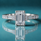 Unique white gold engagement ring with emerald-shaped center diamond, a pair of round diamonds on each side, then a long straight baguette going down the shank, followed by more round diamonds prong-set in channel. There is milgrain surrounding the center diamondn and on both sides of the shank.