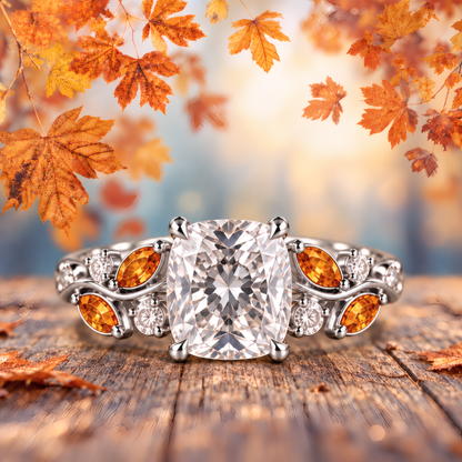 White gold engagement ring with a cushion-cut diamond atop a delicate vine band, woven with fiery amber marquise leaves and twinkling round diamonds for a romantic, nature-inspired look. In a fall-themed environment.