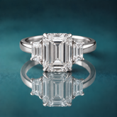 White gold three-stone engagement ring with a large emerald-shaped diamond in the center, flanked by two small trapezoid-shaped diamonds. The shank is slightly rounded and about 3mm wide.