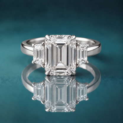 White gold three-stone engagement ring with a large emerald-shaped diamond in the center, flanked by two small trapezoid-shaped diamonds. The shank is slightly rounded and about 3mm wide.