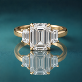 Gold three-stone engagement ring with a large emerald-shaped diamond in the center, flanked by two small trapezoid-shaped diamonds. The shank is slightly rounded and about 3mm wide.
