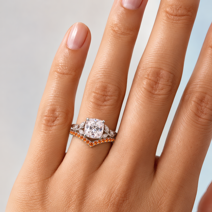 Woman's left ring finger wearing a white gold engagement ring with a cushion-shaped center diamond embraced by a vivid orange curved wedding band and sculpted leaf accents, all the warmth of fall in a single statement piece.
