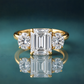 A gold three-stone diamond ring featuring a central emerald-cut diamond flanked by two round-cut diamonds.