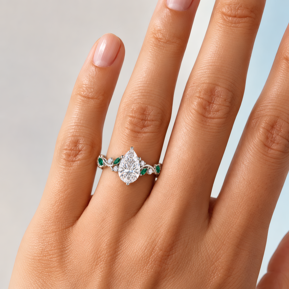 Women's delicate left hand wearing an engagement ring with pear-shaped diamond at its center, cradled by a delicate vine-like shank. Vibrant green emeralds and sparkling round diamonds punctuate the flowing shank.