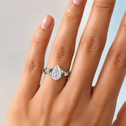 Women's delicate left hand wearing an engagement ring with pear-shaped diamond at its center, cradled by a delicate vine-like shank. Vibrant green emeralds and sparkling round diamonds punctuate the flowing shank.