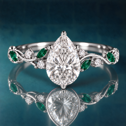 White gold engagement ring with pear-shaped diamond at its center, cradled by a delicate vine-like shank. Vibrant green emeralds and sparkling round diamonds punctuate the flowing shank.